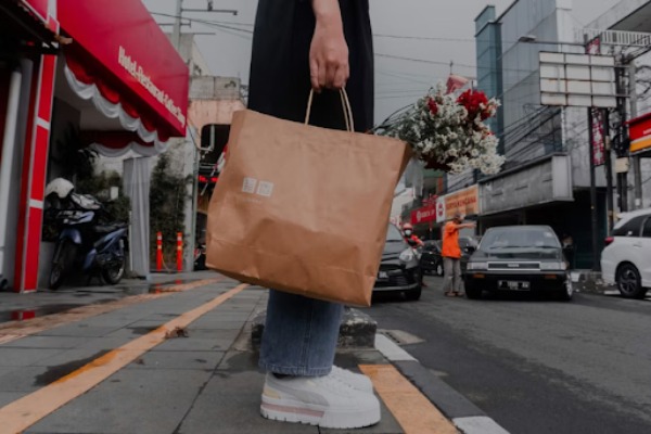 A person in white sneakers standing on the street holding a brown paper bag.
